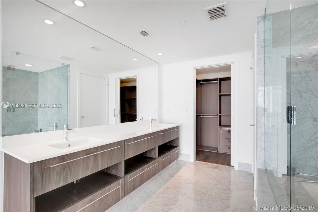 Double vanity with an expansive wall-to-wall mirror, under-mounted sinks, stone-surface countertop, dark wood-finish cabinetry, and glass-enclosed shower with tiled wall surrounds
