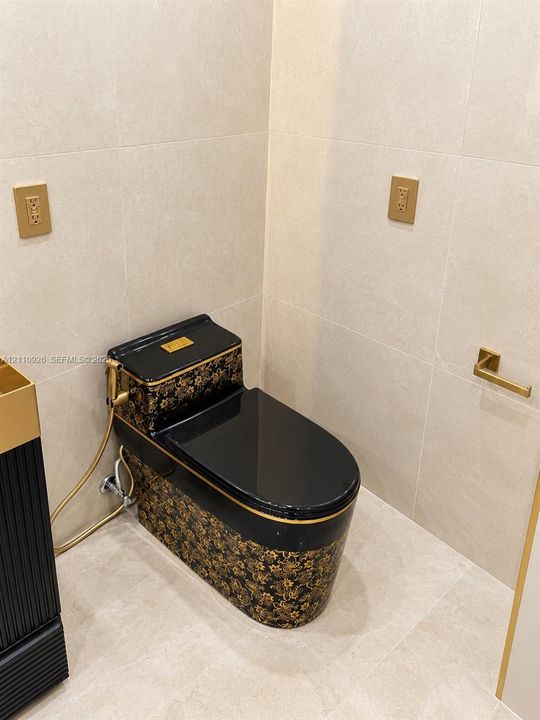 Custom black and gold patterned toilet featuring a matching bidet sprayer, complemented by light-toned large-format wall and floor tile, gold-finish electrical faceplates, and a wall-mounted gold-finish paper holder