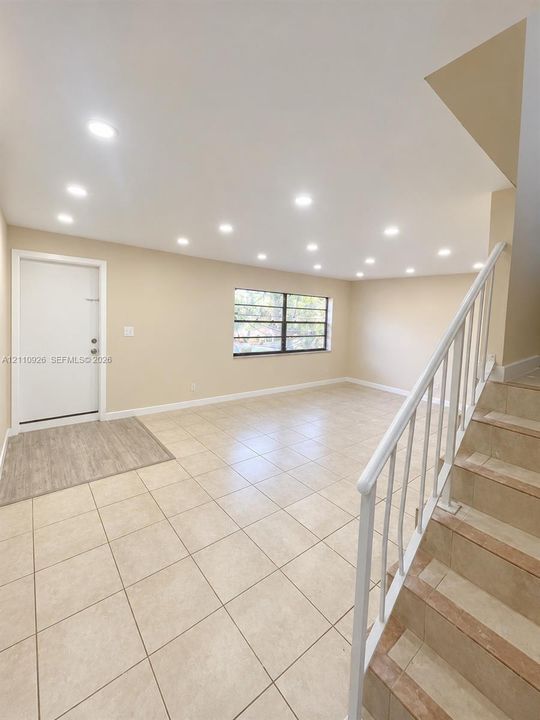 Open-concept interior featuring light-toned tile flooring, recessed ceiling lighting, a multi-pane window, and a staircase with a white metal railing