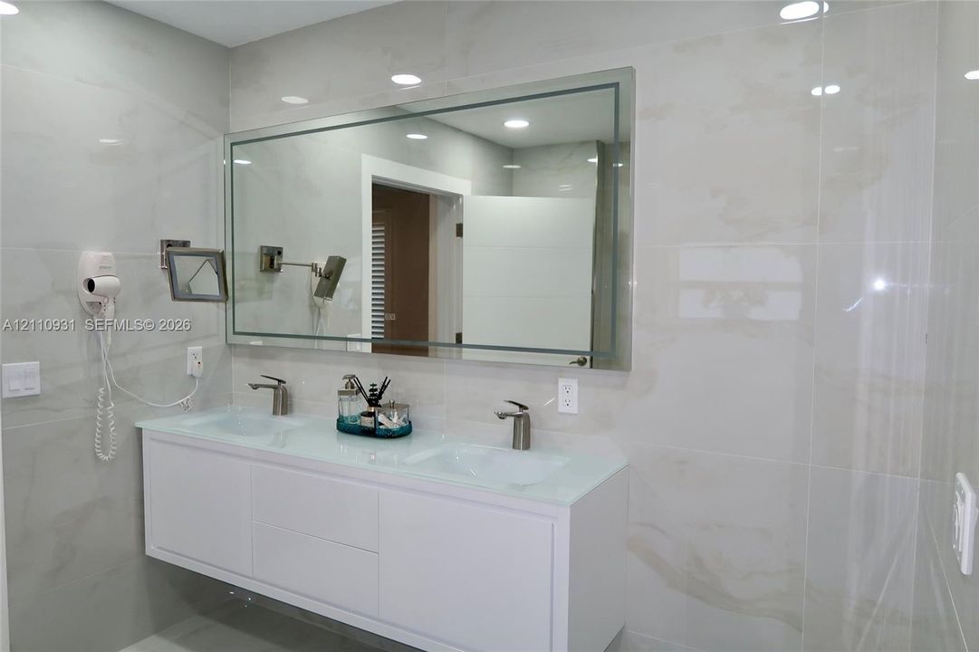 Wall-mounted white dual-sink vanity featuring a glass countertop, large rectangular mirror with integrated lighting, wall-mounted adjustable magnification mirror, and large-format neutral-toned tile wall surfacing