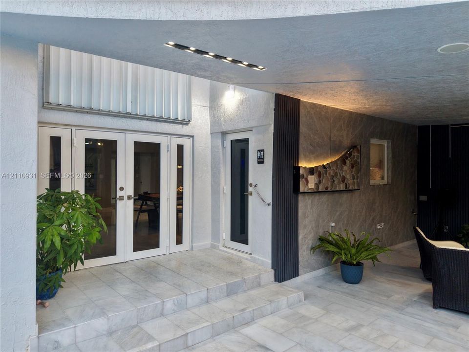 Covered exterior entryway featuring stone-tiled flooring, double glass-panel doors, accent stone wall cladding with integrated lighting, and vertical slat detailing