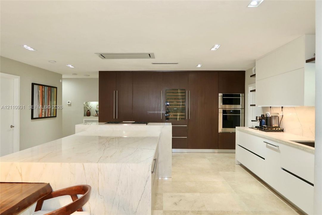 Kitchen featuring stone-topped waterfall island, dark wood-finish cabinetry, built-in wine refrigeration, integrated wall ovens, and stone flooring