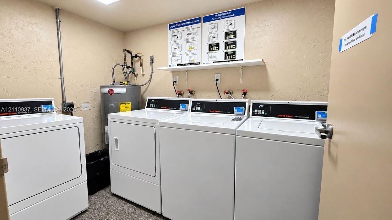 Laundry room featuring multiple commercial-grade top-load washing machines, a matching dryer, a wall-mounted shelving unit, and an integrated water heater