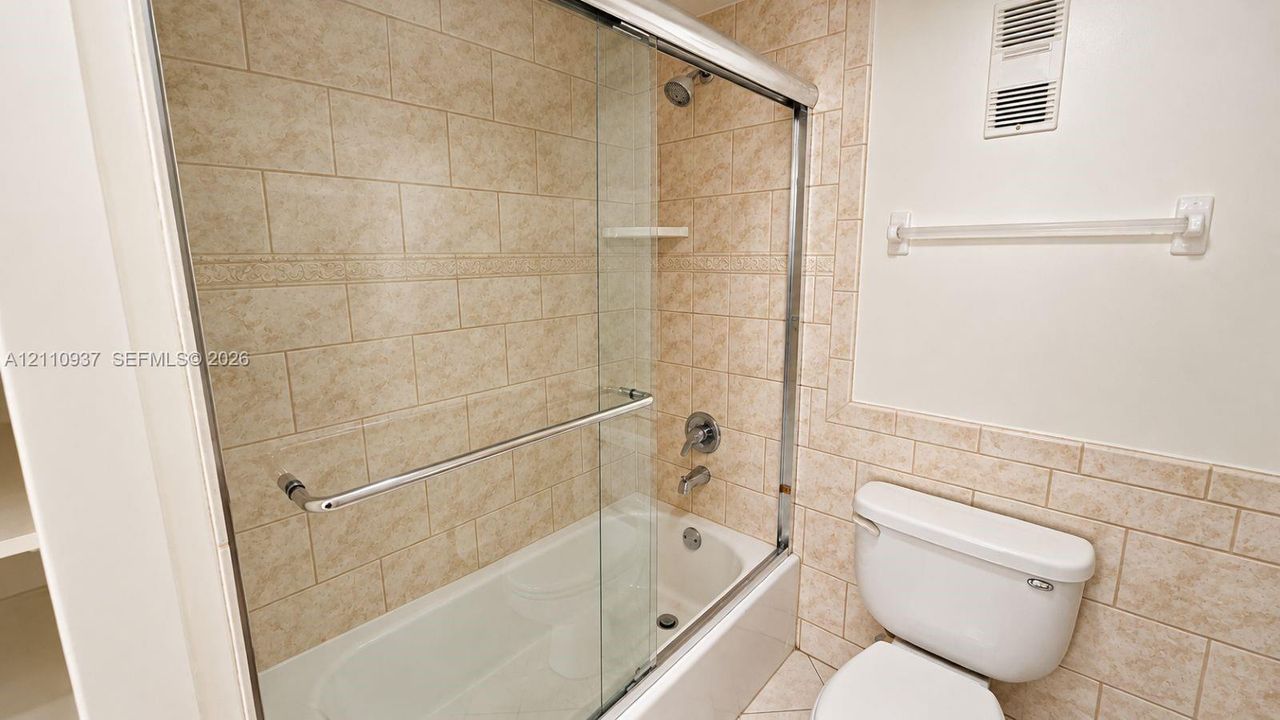 Full-height tub enclosure with sliding glass door, neutral stone-finish ceramic tile surround, integrated shelving, and polished chrome hardware