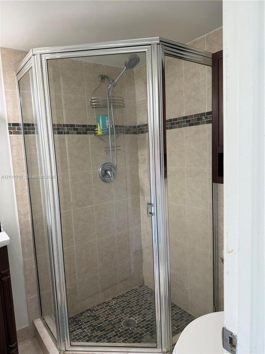 Corner shower enclosure featuring a metal frame, tile wall surround, stone-tiled floor, decorative glass tile accent strip, and a chrome hand-held shower fixture