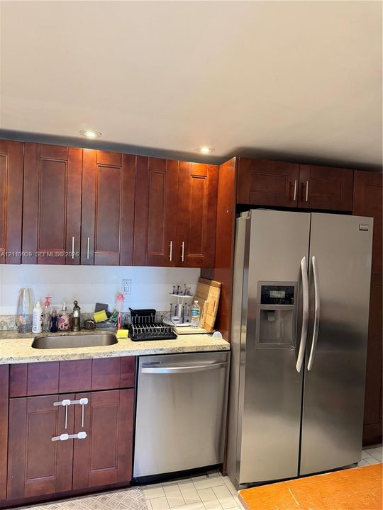 Kitchen area featuring dark-stained wood cabinetry, stone-finish countertops, stainless steel side-by-side refrigerator, under-mount stainless steel sink, and stainless steel dishwasher
