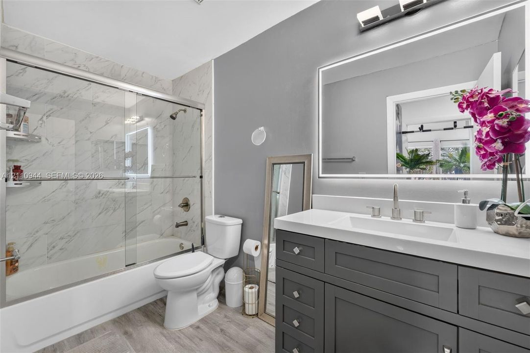 Shaker-style vanity with a solid-surface countertop and integrated sink, illuminated LED mirror, tub-shower combination with sliding glass enclosure, stone-patterned tile shower surround, and wood-finish flooring
