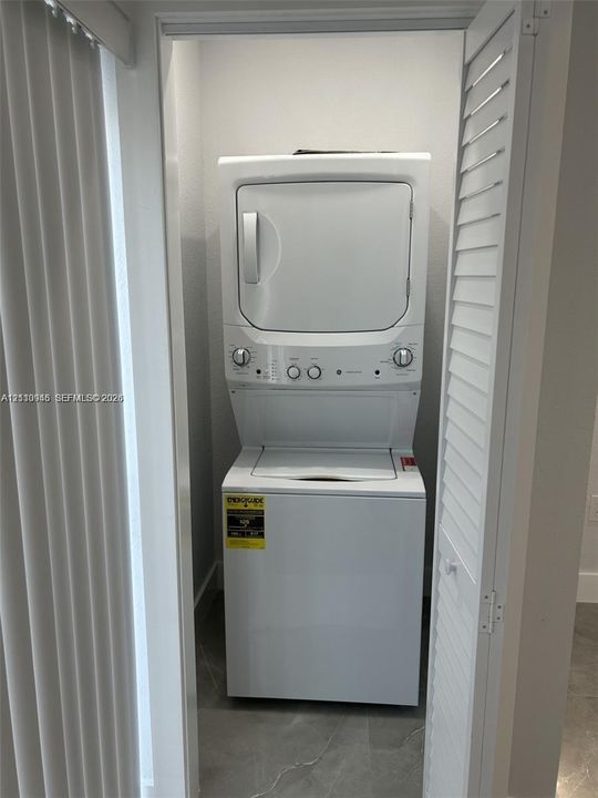 Stacked white laundry unit enclosed by louvered bi-fold doors, featuring tile flooring and vertical blind hardware