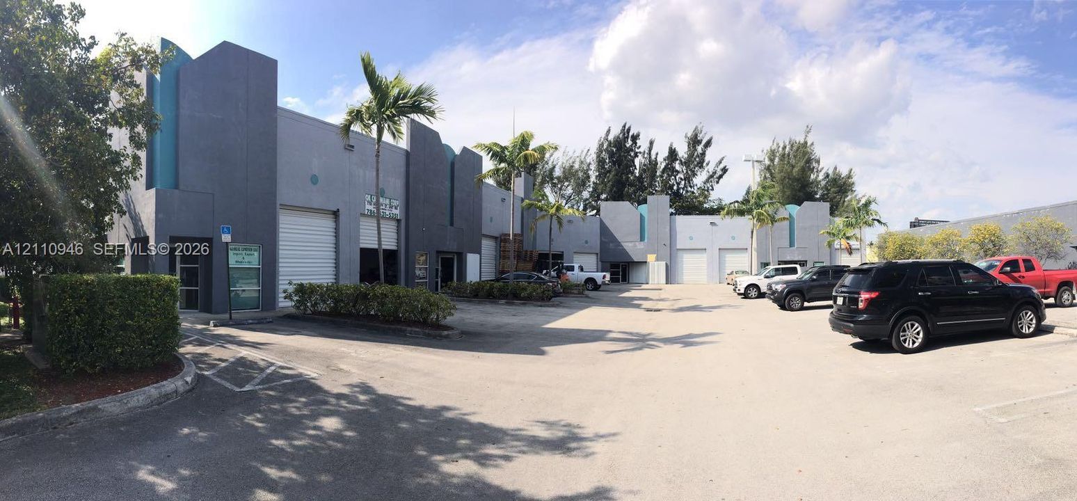 Commercial building exterior featuring a grey stucco facade, multiple overhead roll-up bay doors, private asphalt parking area, and mature palm tree landscaping