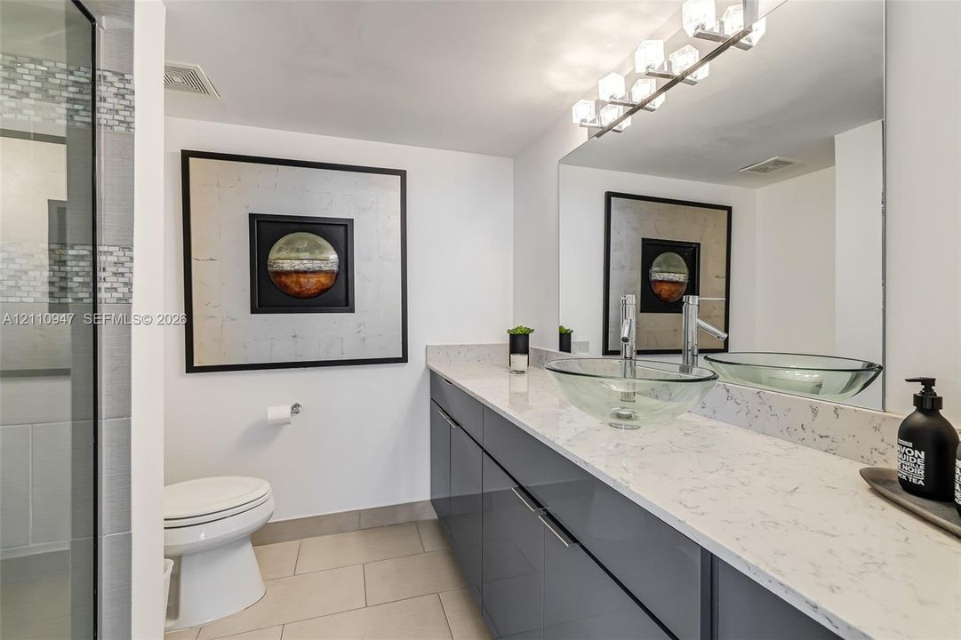 Modern dual-vanity featuring gloss-finish cabinetry, stone-patterned countertops, glass vessel sinks, and an integrated full-width mirror with multi-bulb light fixture