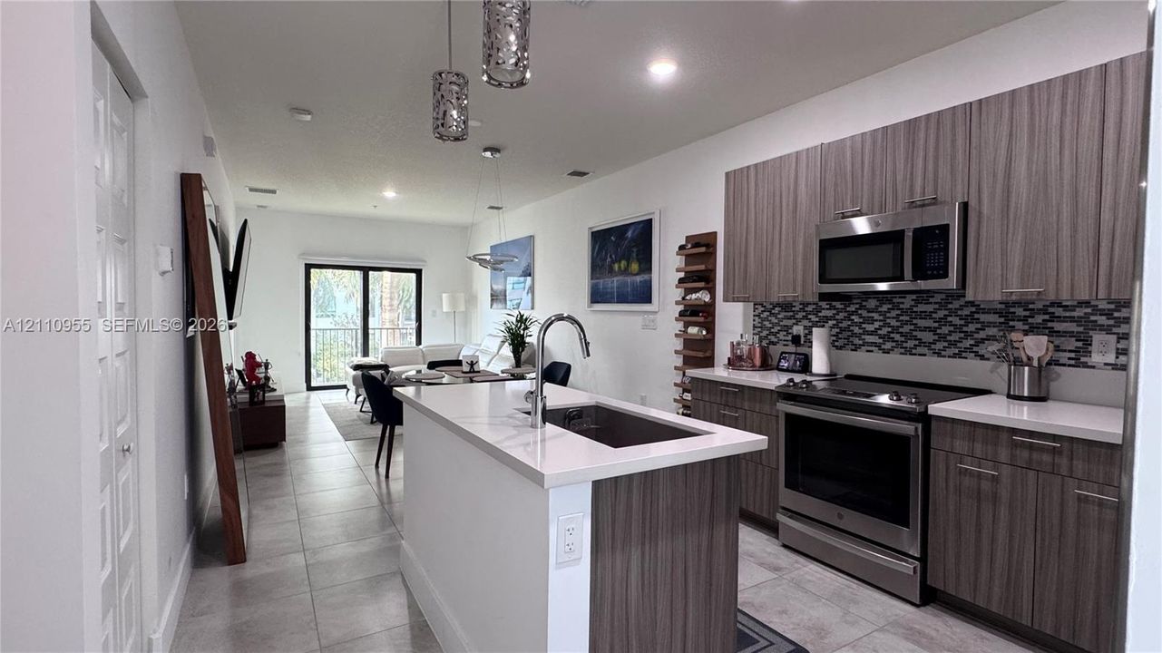 Kitchen featuring flat-panel wood-finish cabinetry, light-toned stone countertops, a multi-tonal mosaic tile backsplash, stainless steel appliances, and a central island with an integrated sink and gooseneck faucet
