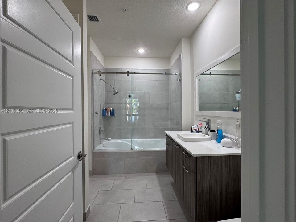 Bathroom featuring a vanity with wood-finish cabinetry, white countertop, rectangular sink, full-width mirror, and a tub-shower combination with a sliding glass door and large-format tile surround