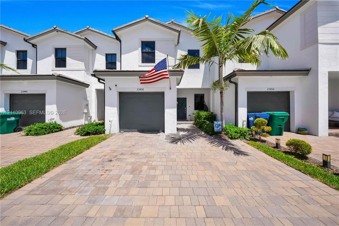 Multi-story townhouse exterior featuring a white stucco facade, paver driveway, dark-toned garage doors, and a decorative palm tree