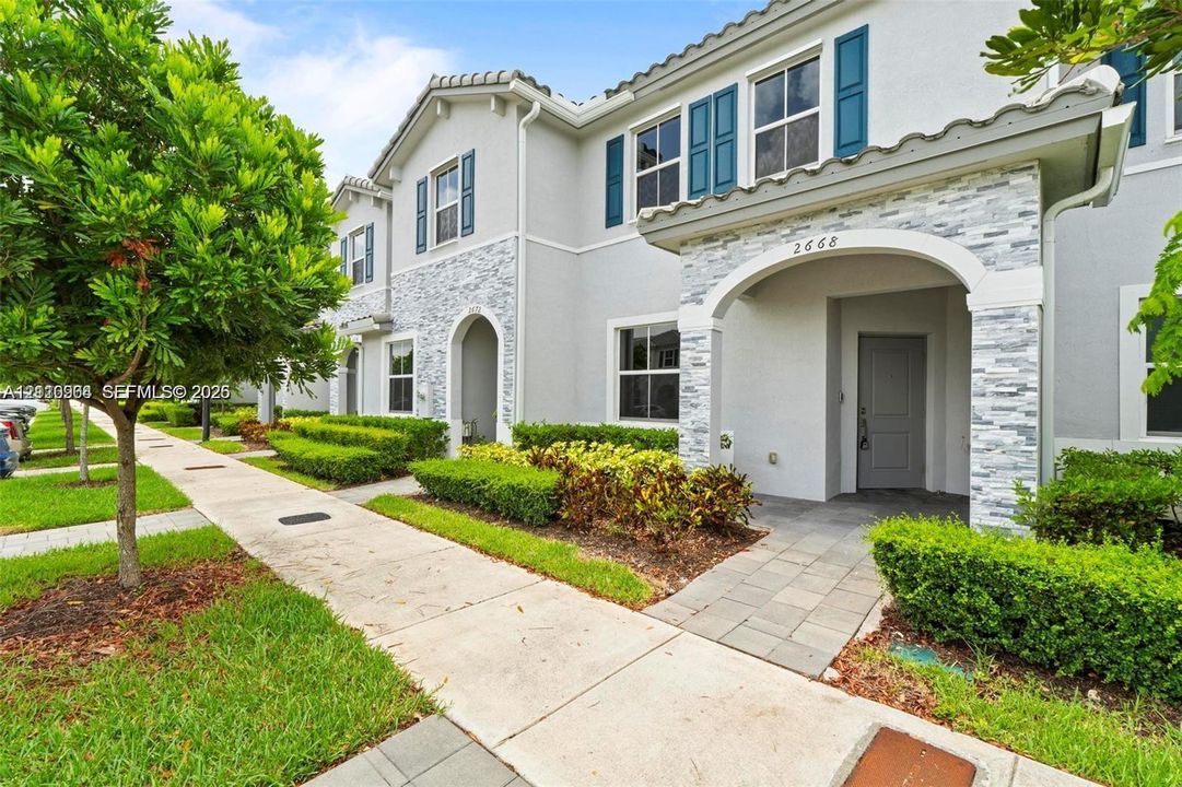 Two-story stucco exterior featuring stacked stone veneer accents, arched covered entryways, blue shutter detailing, and paved walkways