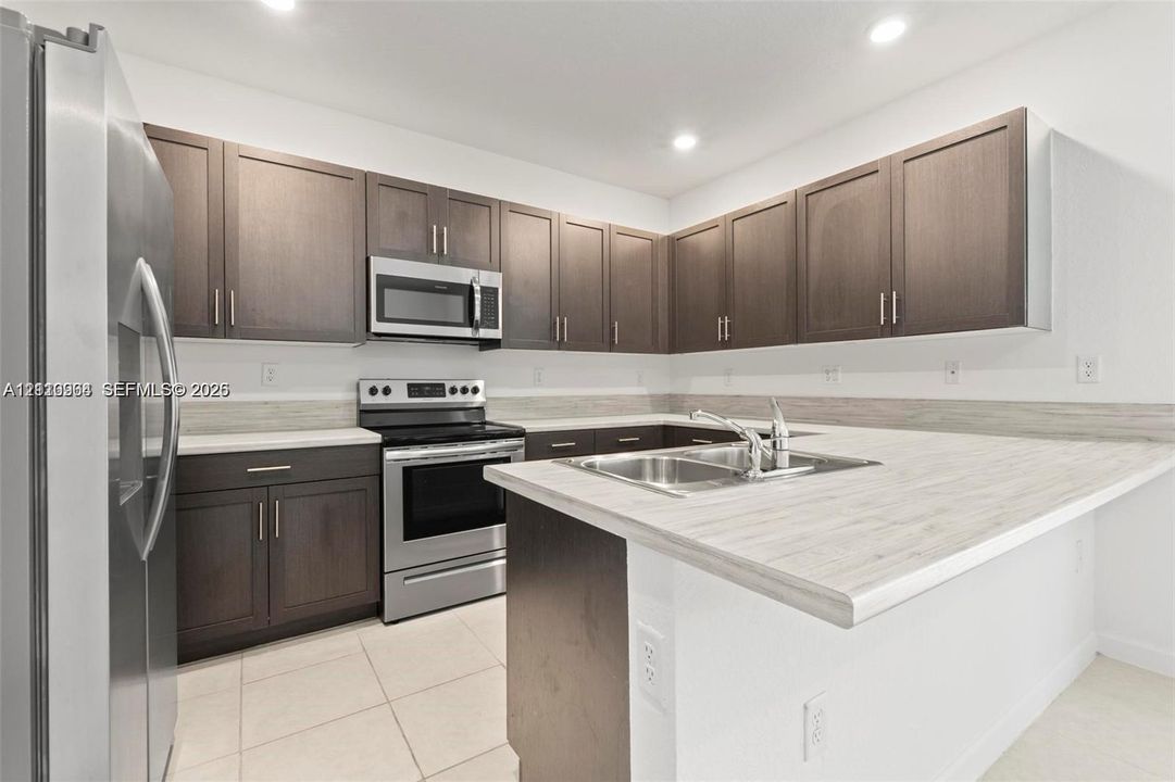 Kitchen featuring dark wood-finish cabinetry, light-toned laminate countertops, stainless steel appliances, a double-basin stainless steel sink, and tile flooring