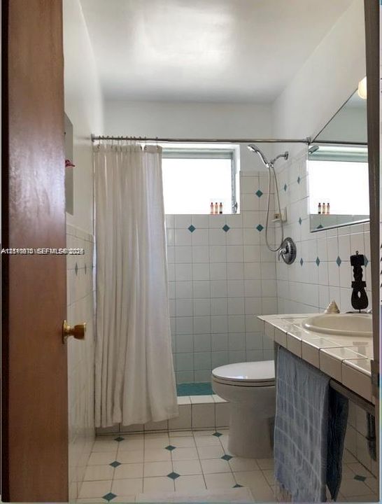 Bathroom featuring white tiled walls with diamond-shaped accent inlays, tiled countertop with an integrated sink, shower stall with a handheld showerhead, and a wall-mounted mirror