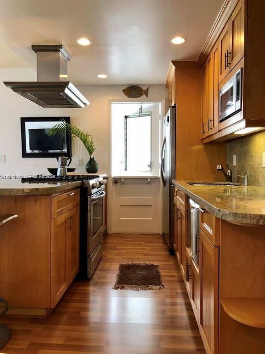 Kitchen featuring wood cabinetry, stone countertops, stainless steel appliances, a ceiling-mounted range hood, and wood-finish flooring