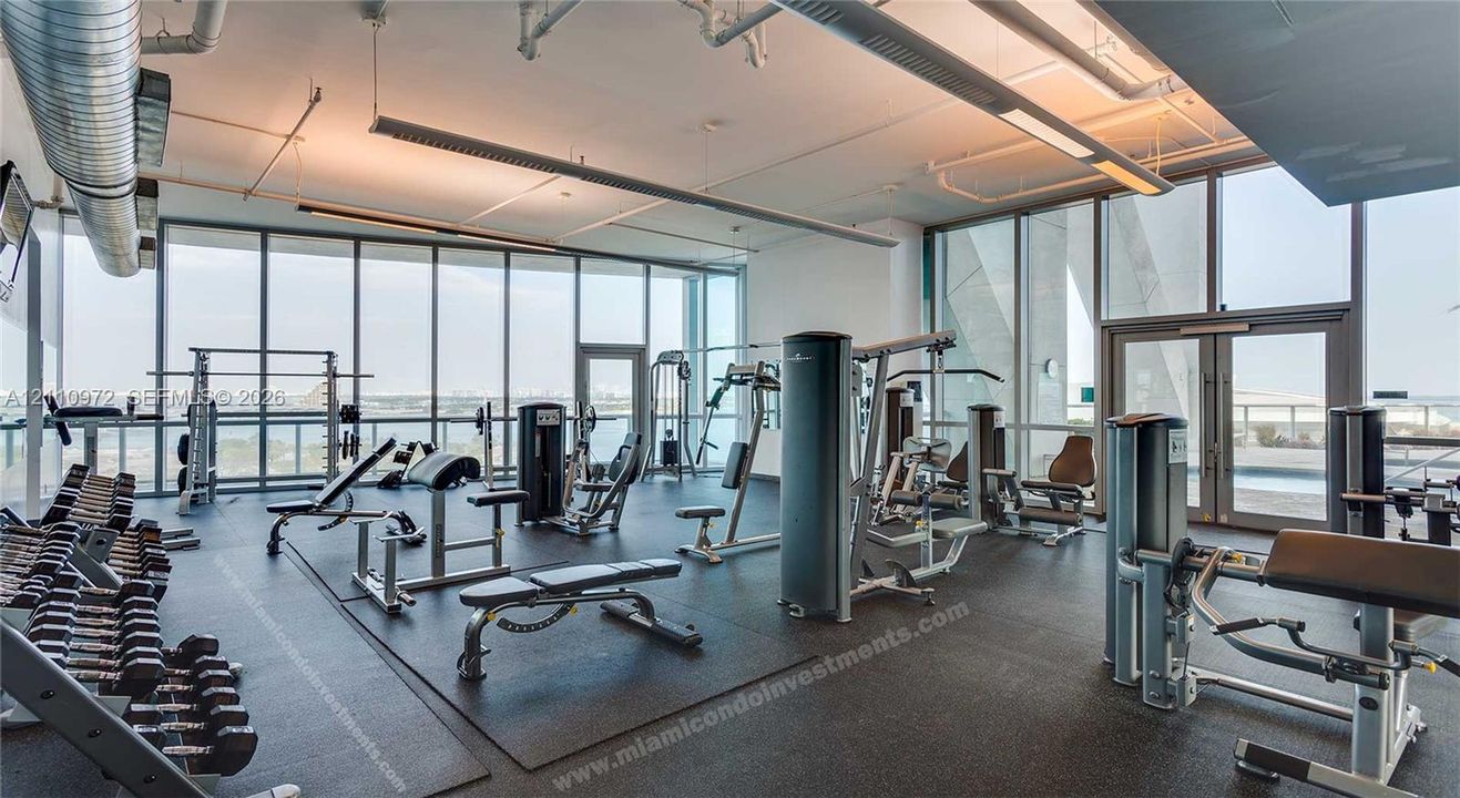 Fitness center featuring floor-to-ceiling windows, exposed industrial ductwork, linear pendant lighting, and rubberized flooring