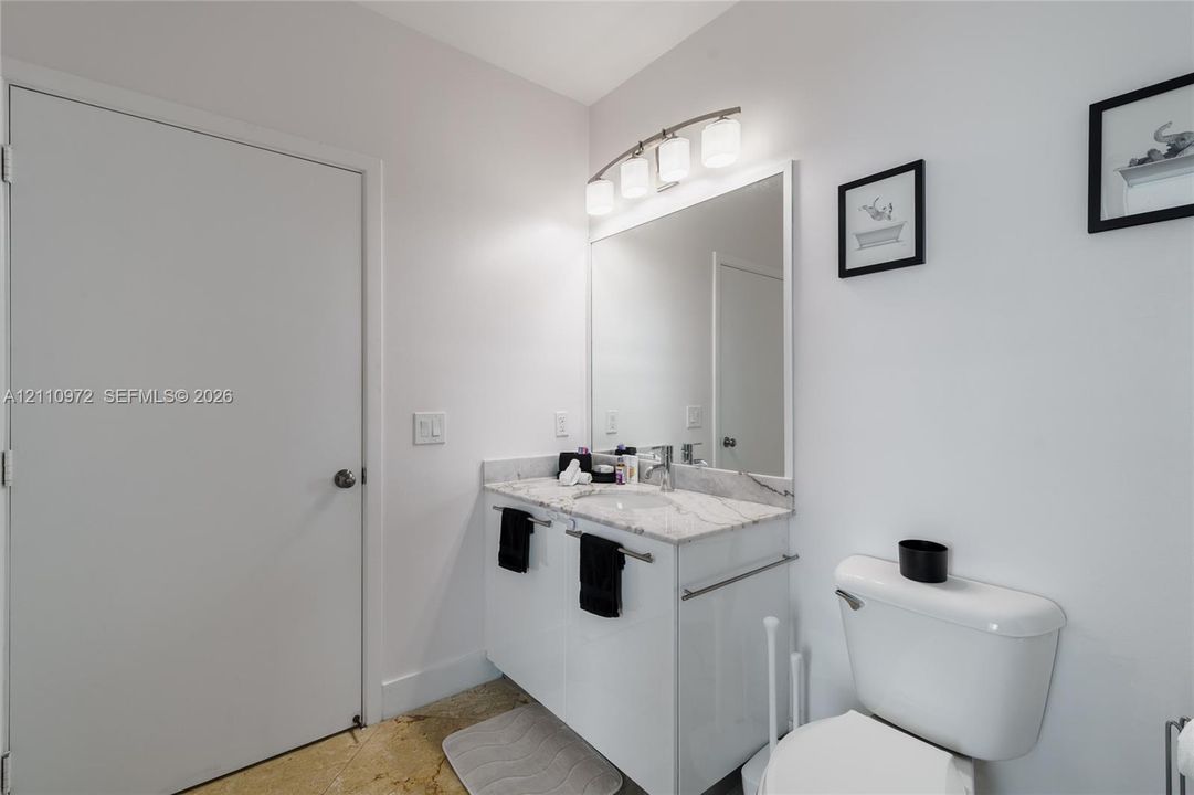White vanity with a stone countertop, integrated undermount sink, wall-mounted vanity mirror, and three-light chrome fixture
