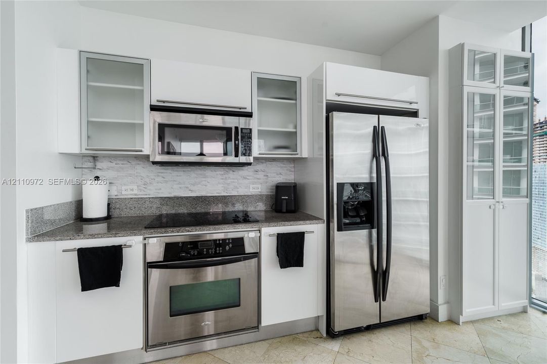 Kitchen featuring high-gloss white cabinetry, stone-finish countertops, tile backsplash, built-in stainless steel appliances, and glass-front display cabinetry