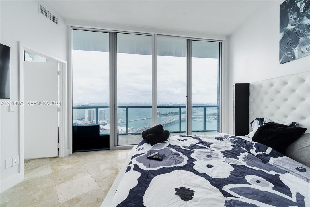 Floor-to-ceiling glass paneling, sliding glass door access to a balcony with a metal railing, stone-finish flooring, and expansive water and urban skyline scenery