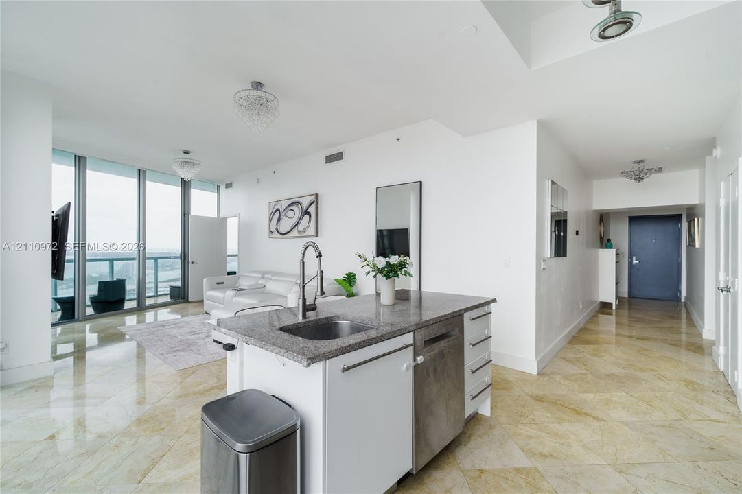 Kitchen island featuring stone-finish countertops, stainless steel undermount sink, integrated dishwasher, and built-in cabinetry, complemented by floor-to-ceiling windows and polished stone-finish flooring