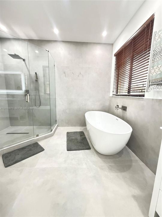 Freestanding oval soaking tub with wall-mounted faucet, walk-in glass-enclosed shower, large-format tile wall and floor surfaces, and recessed ceiling lighting