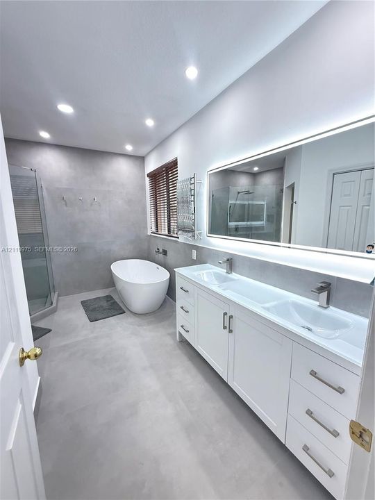 Dual-vanity cabinet with integrated sinks and metallic hardware, expansive backlit mirror, freestanding soaking tub, enclosed glass shower, and large-format tile flooring