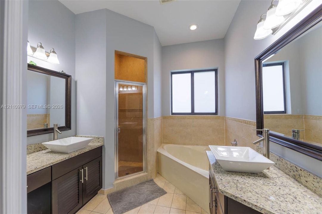 Dual vanities featuring rectangular vessel sinks, stone-finish countertops, dark wood-finish cabinetry, and framed mirrors, complemented by a corner soaking tub and a separate glass-enclosed shower