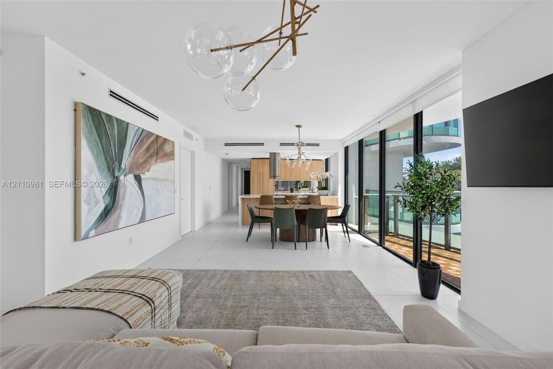 Open-concept interior featuring large-format tile flooring, floor-to-ceiling glass panel walls, custom light-toned wood cabinetry, and an architectural multi-sphere pendant fixture