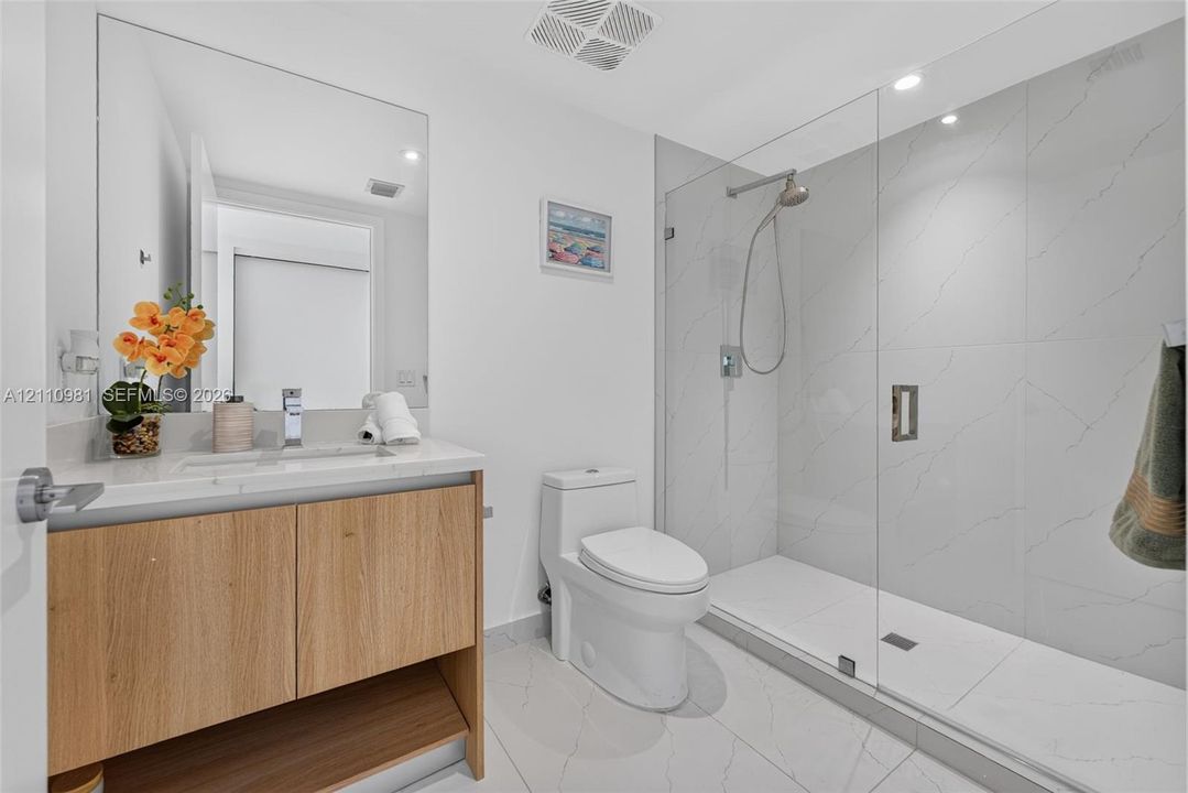 Wood-finish vanity with a stone-surfaced countertop, glass-enclosed shower featuring large-format tile, and a wall-to-wall mirror