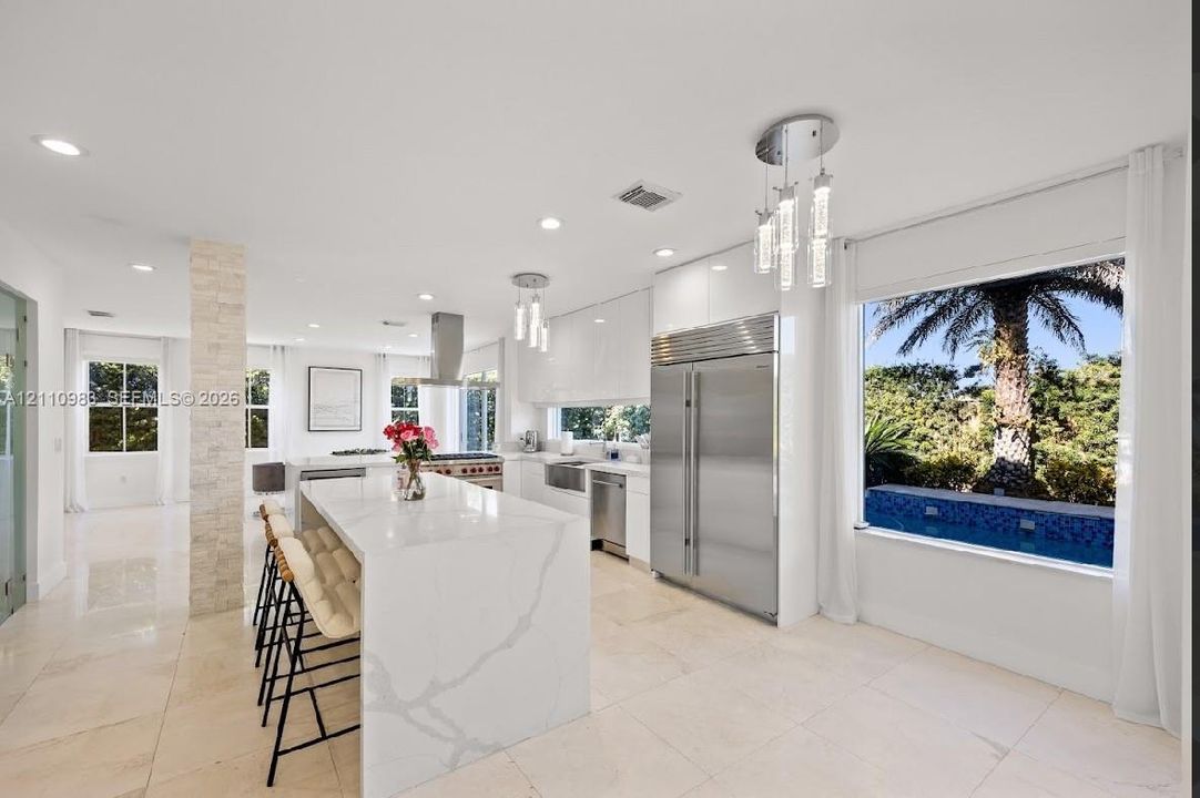 Contemporary kitchen featuring an oversized center island with waterfall edge, full-height high-gloss cabinetry, professional-grade stainless steel refrigerator, recessed lighting, and tile flooring