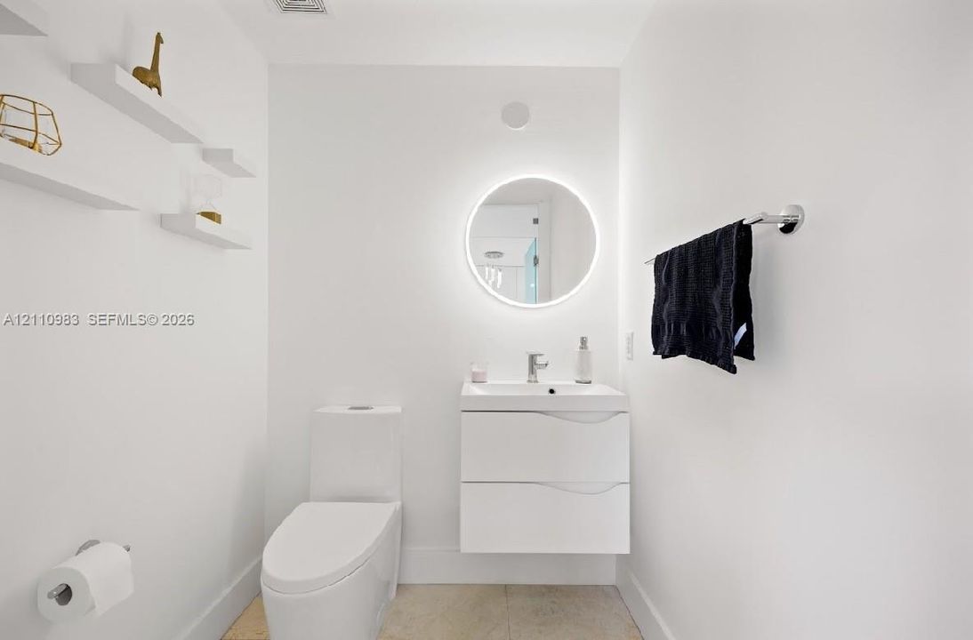 Modern floating vanity with integrated sink, backlit circular wall mirror, wall-mounted shelving, and polished-finish tile flooring