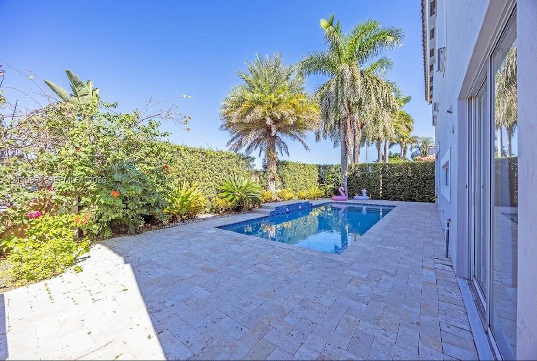 Outdoor patio featuring natural stone pavers, a rectangular swimming pool with a built-in water feature, and mature perimeter landscaping including tall palm trees