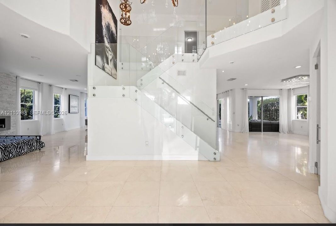Grand foyer featuring polished light-toned stone flooring, a sweeping staircase with a structural glass railing, recessed ceiling lighting, and multiple large-scale windows
