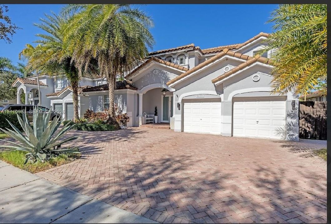 Multi-gabled exterior featuring a neutral stucco facade, clay tile roofing, paved brick driveway, dual garage doors, and arched entryway