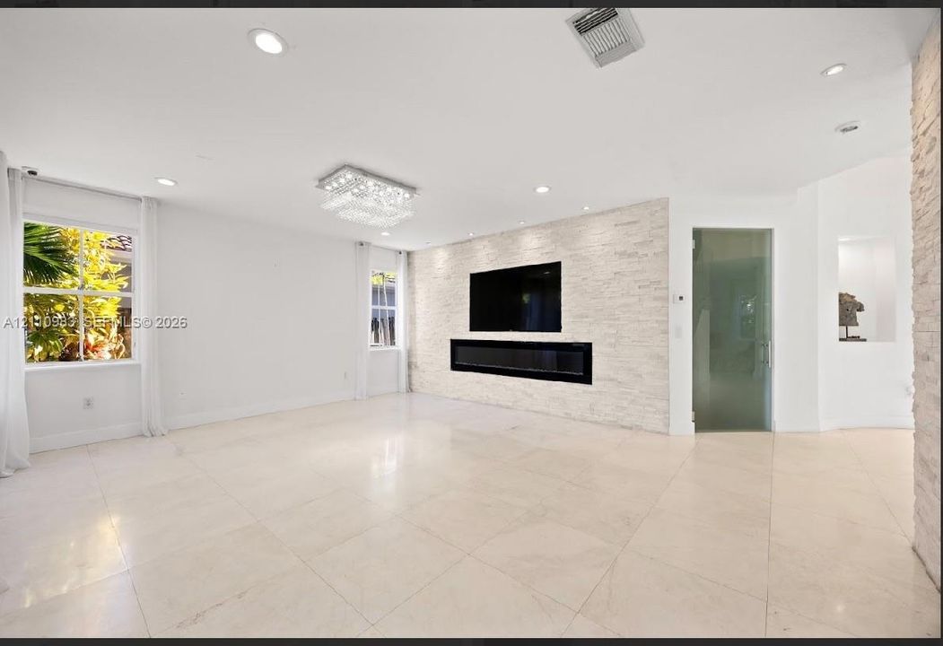 Living space featuring large-format tile flooring, textured stone accent wall with integrated linear electric fireplace, recessed ceiling lighting, a tiered crystal chandelier, and a glass-paneled door