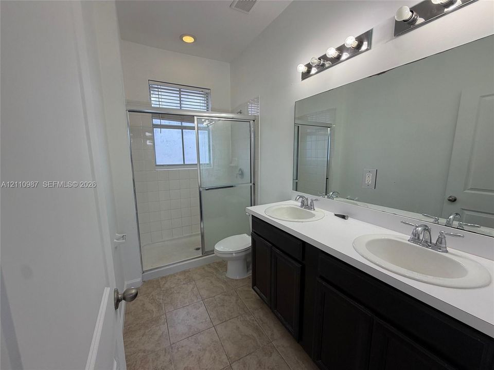 Dual-sink vanity with dark cabinetry and a solid-surface countertop, full-width vanity mirror, tile flooring, and a walk-in shower with a glass enclosure