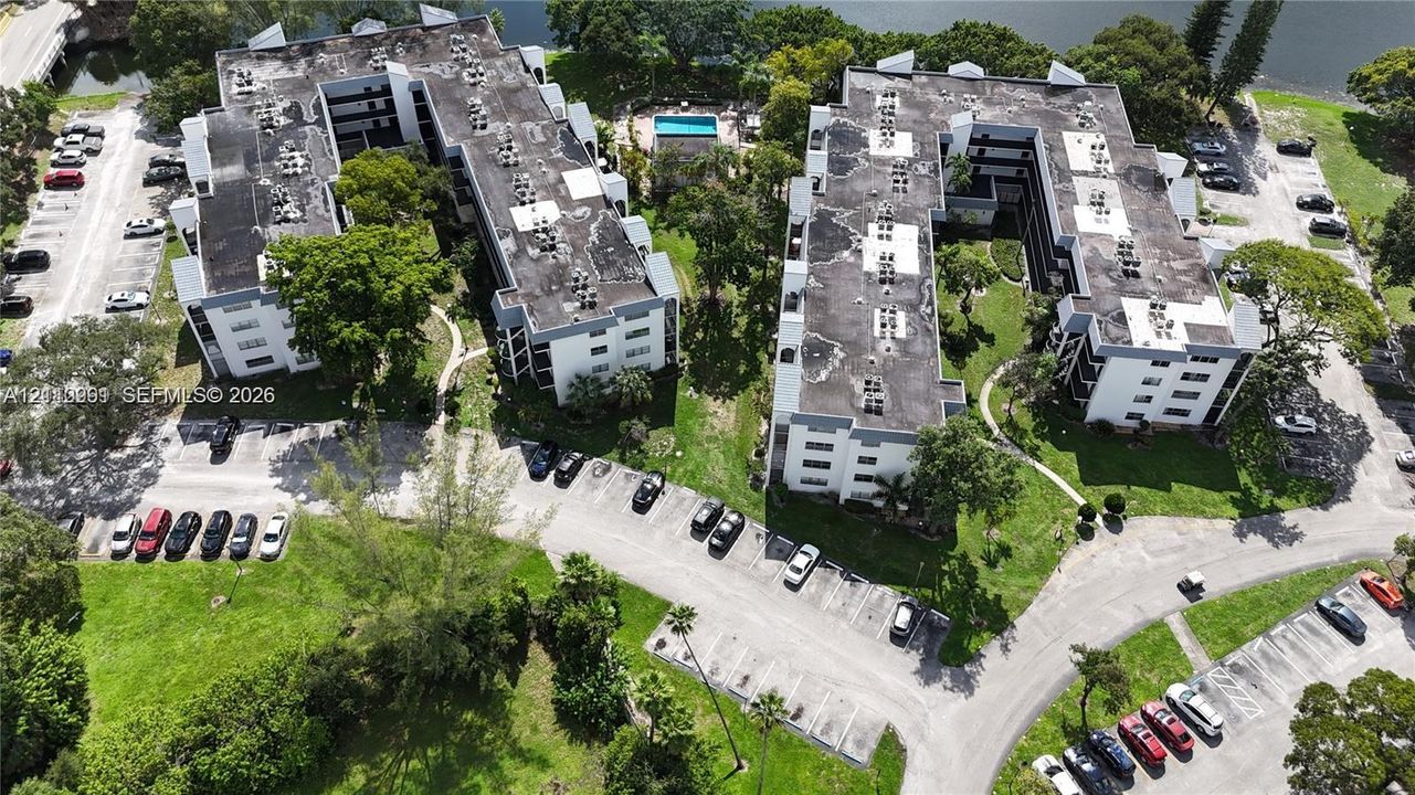 Aerial perspective of a multi-story residential complex featuring paved surface parking, central courtyard landscaping, and an outdoor swimming pool
