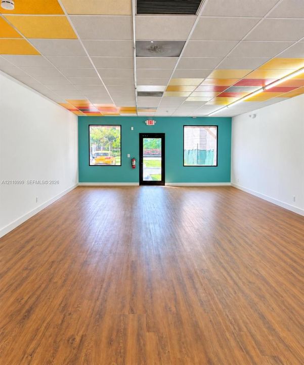 Open-plan interior space featuring wood-finish flooring, drop-ceiling grid with multi-colored acoustic panels, and two double-pane windows flanking a central glass-panel entry door