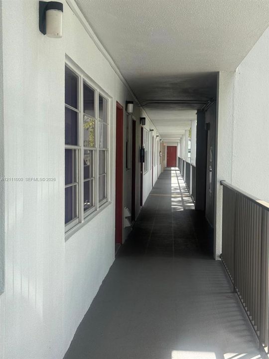 Covered exterior walkway featuring painted stucco walls, multi-pane windows, metal railings, and wall-mounted light fixtures