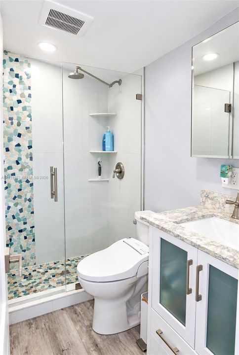Walk-in shower with a glass enclosure, pebble-stone flooring, decorative mosaic wall tile, and built-in corner shelving, accompanied by a vanity with frosted cabinet doors and stone-finish countertop