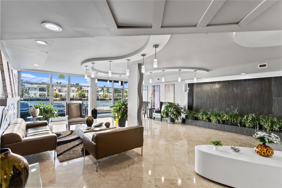 Expansive open-concept space featuring polished stone flooring, a decorative stone-tiled water feature, curvilinear coffered ceiling accents, and floor-to-ceiling glass windows with water frontage