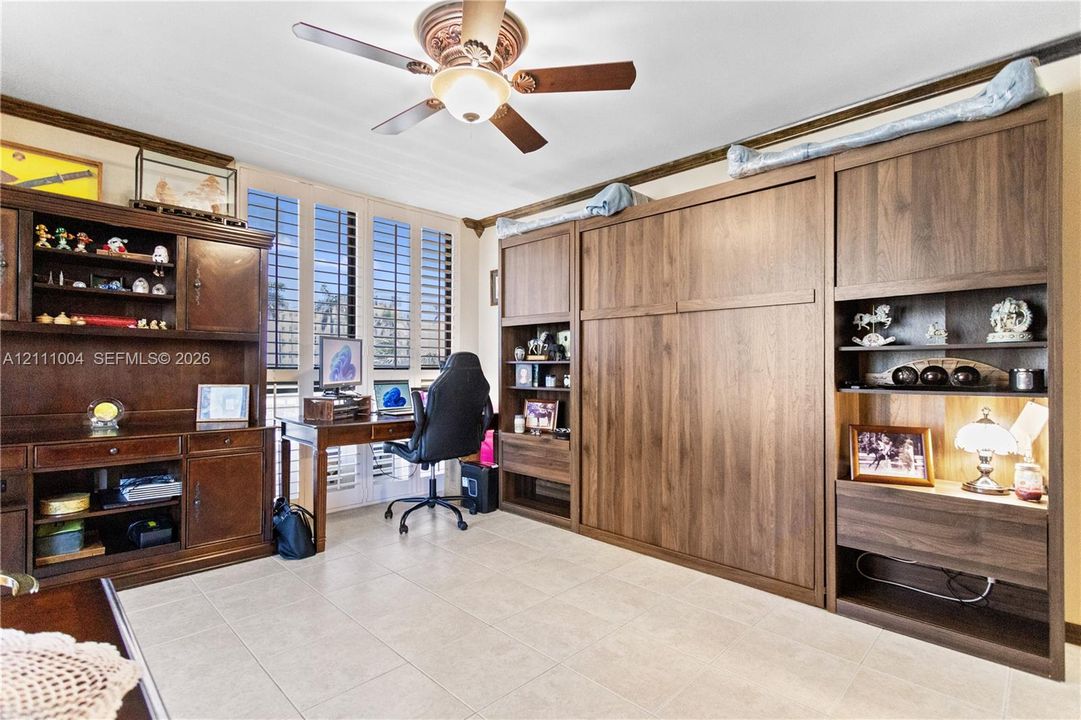 Custom wood-finish cabinetry featuring integrated desk space and built-in shelving, complemented by a ceiling fan and plantation shutters