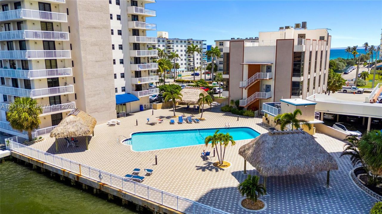 Outdoor recreational area featuring a swimming pool, paved sundeck, and multiple thatched-roof gazebos