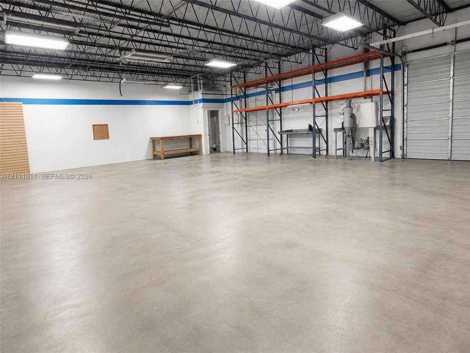 Open-span warehouse interior featuring polished concrete flooring, exposed overhead trusses, high-intensity industrial lighting, and a metal roll-up door