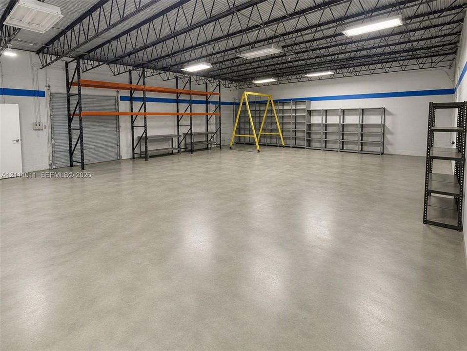 Industrial warehouse interior featuring exposed steel ceiling trusses, polished concrete flooring, overhead fluorescent lighting, and extensive built-in steel shelving units