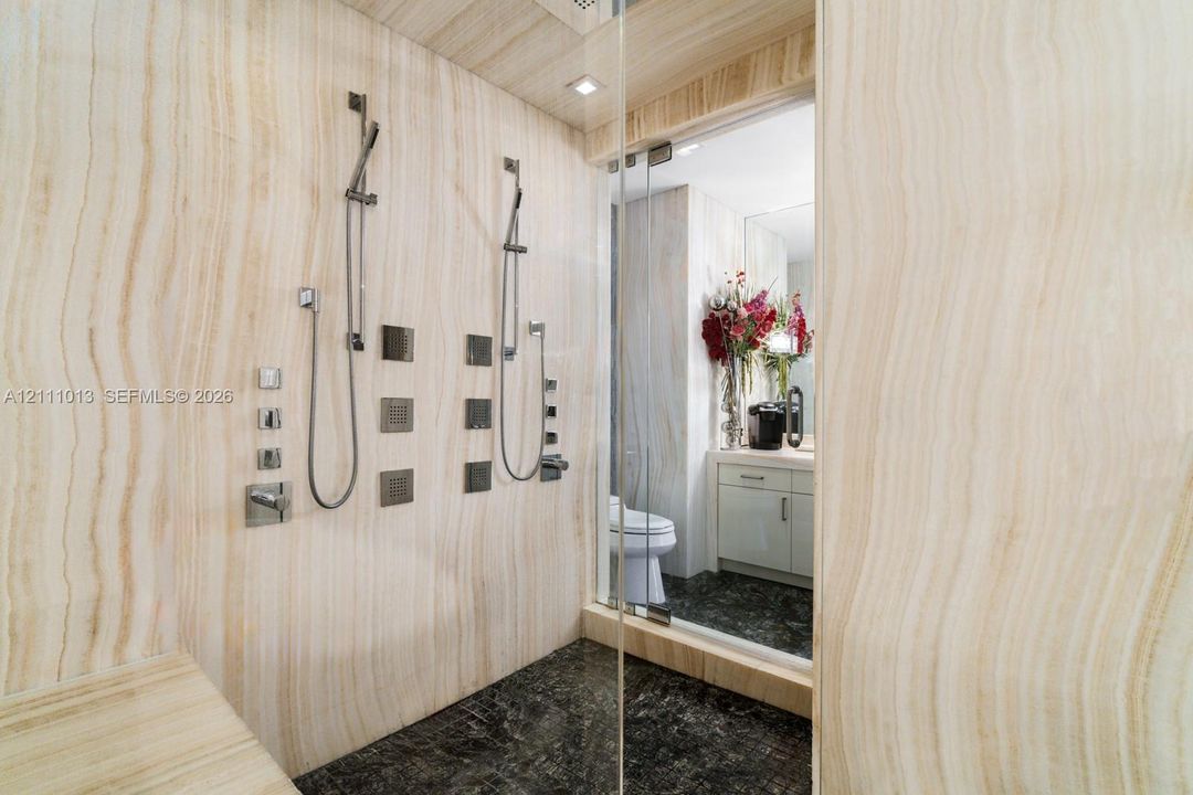 Walk-in shower featuring full-height natural stone wall cladding, dual handheld shower heads with integrated body sprayers, a built-in stone bench, and glass enclosure
