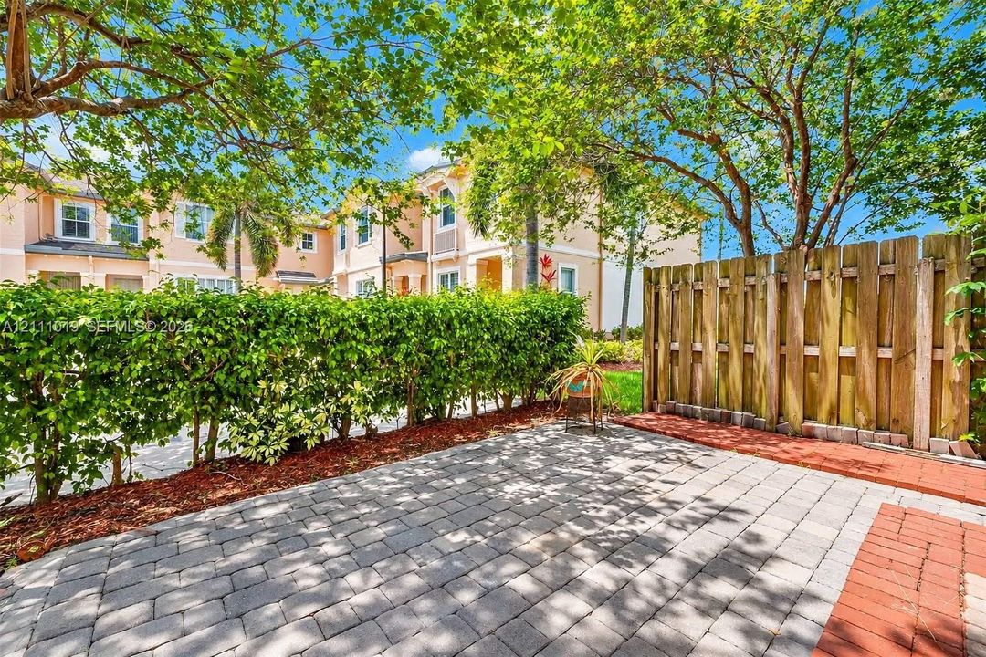 Exterior patio area featuring concrete paver flooring, red brick border detailing, and wood privacy fencing, bordered by established hedges and mature trees