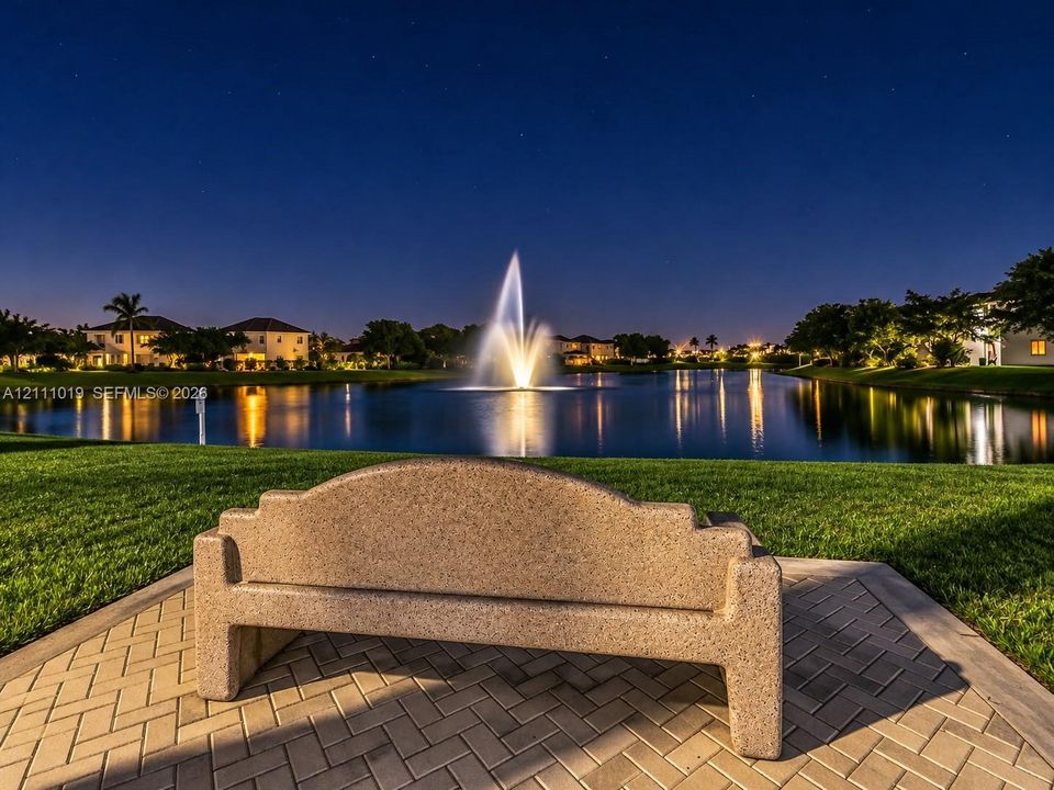 Concrete bench situated on a herringbone-patterned paver patio overlooking a pond with a central water fountain and surrounding manicured turf